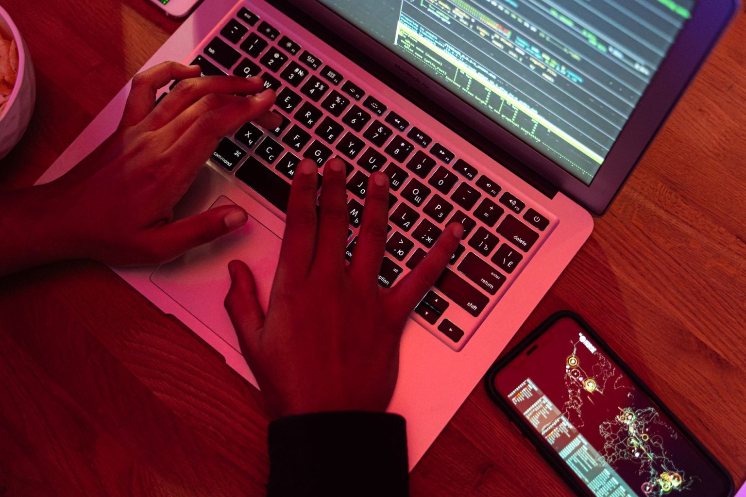 Mãos digitam em um notebook com códigos na tela, ao lado de um smartphone, em um ambiente escuro.