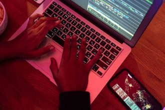 Mãos digitam em um notebook com códigos na tela, ao lado de um smartphone, em um ambiente escuro.