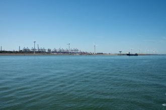 Parque eólico no mar próximo a um porto, com embarcações e atividade marítima na costa.