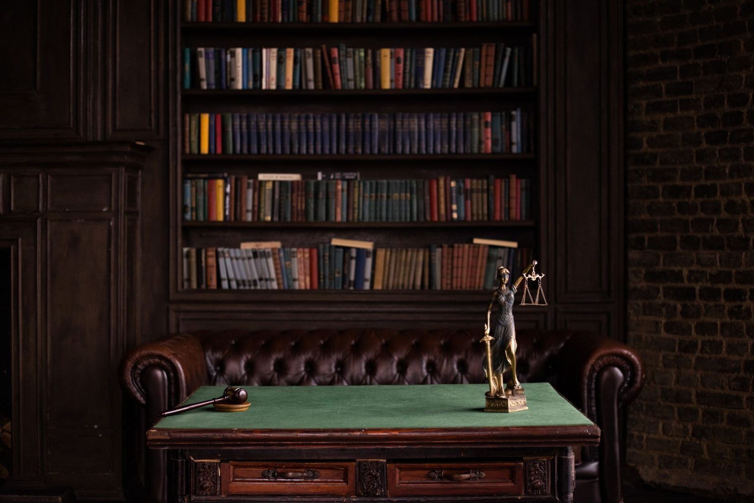 Sala de estudos com estantes de livros jurídicos, um martelo de juiz e uma representação da Justiça sobre uma mesa verde.