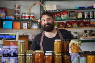 Comerciante sorrindo atrás de prateleiras com potes de conservas artesanais em sua loja.